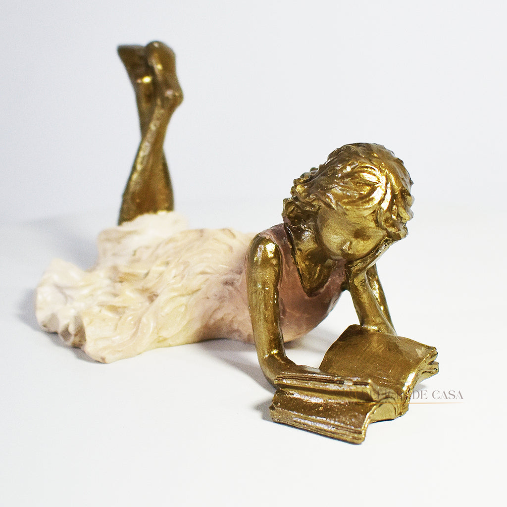Escultura decorativa de menina lendo, deitada com as pernas cruzadas, feita em resina dourada com vestido em tons claros. Decoração para apaixonados por livros.