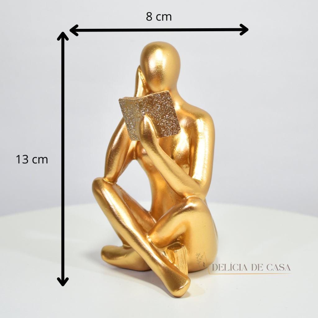 Escultura decorativa dourada de mulher lendo, sentada sobre livros, com detalhes em glitter, ideal para decoração moderna e sofisticada