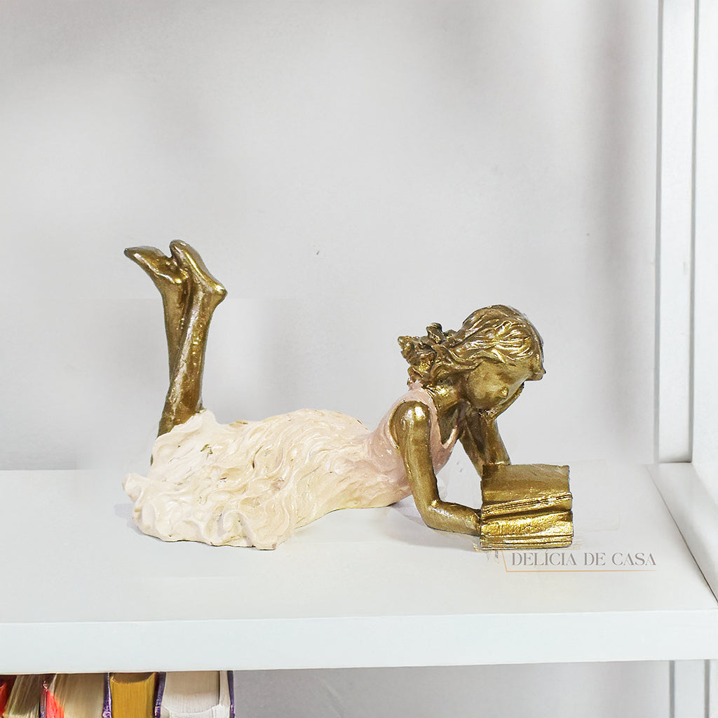 Escultura decorativa de menina lendo, deitada com as pernas cruzadas, feita em resina dourada com vestido em tons claros. Decoração para apaixonados por livros.