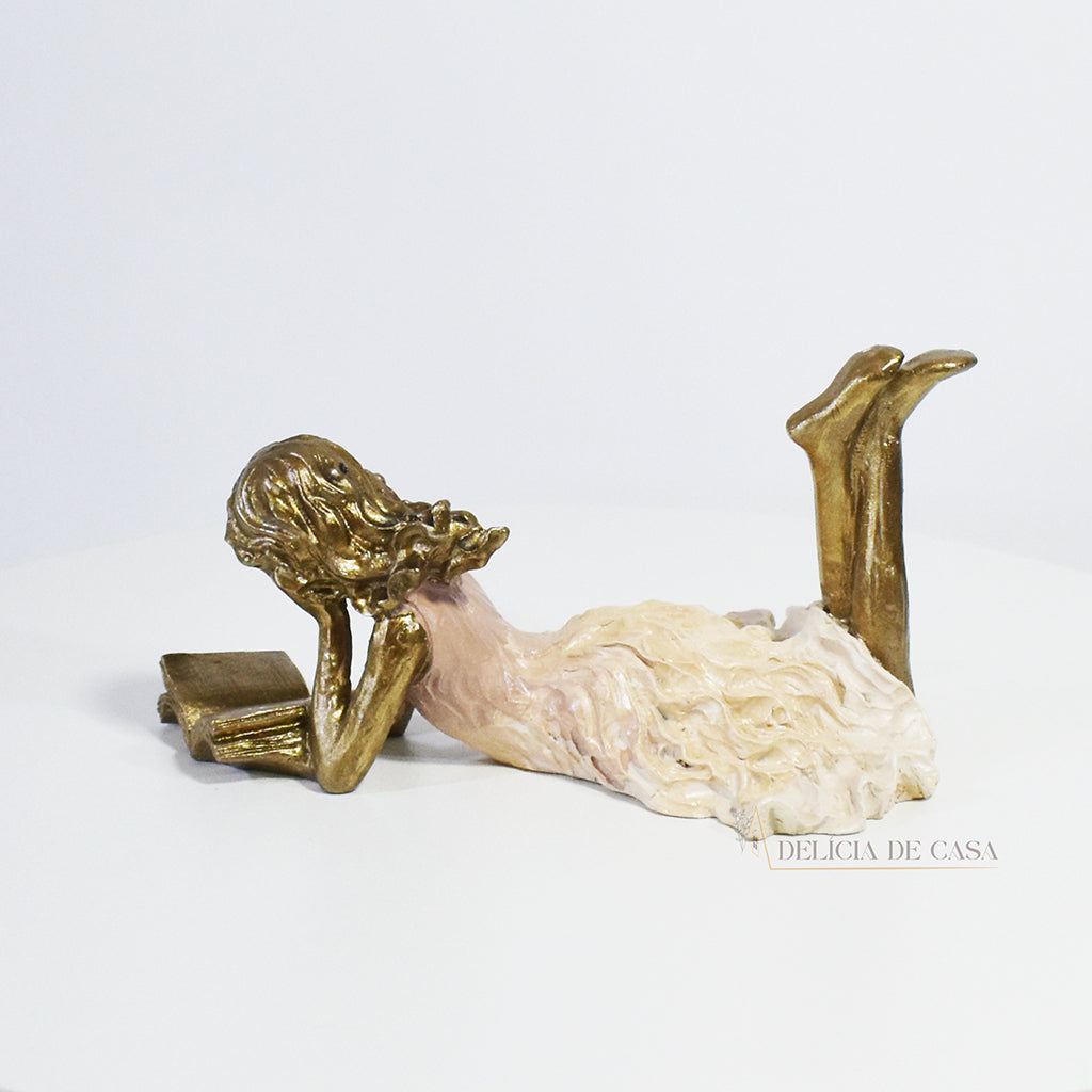 Escultura decorativa de menina lendo, deitada com as pernas cruzadas, feita em resina dourada com vestido em tons claros. Decoração para apaixonados por livros.