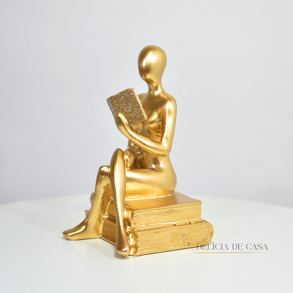 Escultura decorativa dourada de mulher lendo, sentada sobre livros, com detalhes em glitter, ideal para decoração moderna e sofisticada