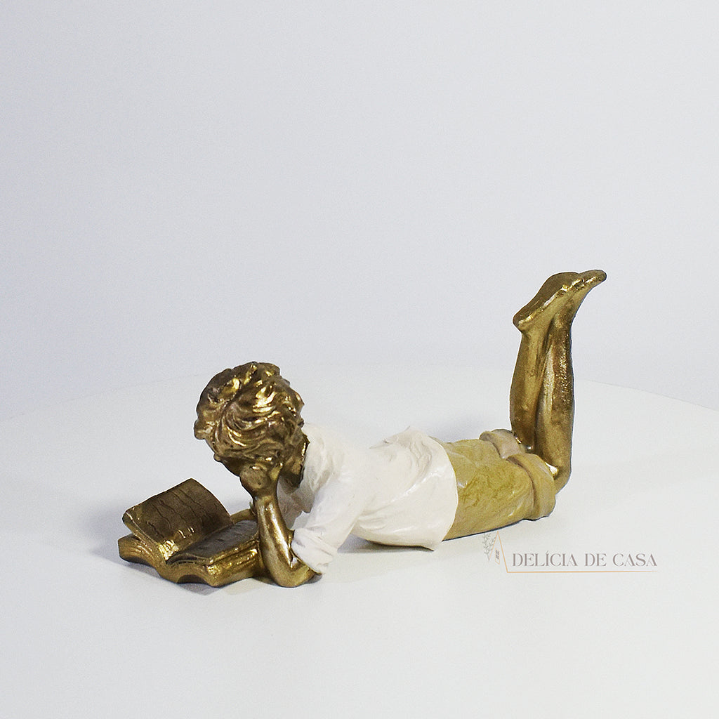 Escultura decorativa de menino lendo, deitada com as pernas cruzadas, feita em resina dourada com vestido em tons claros. Decoração para apaixonados por livros.