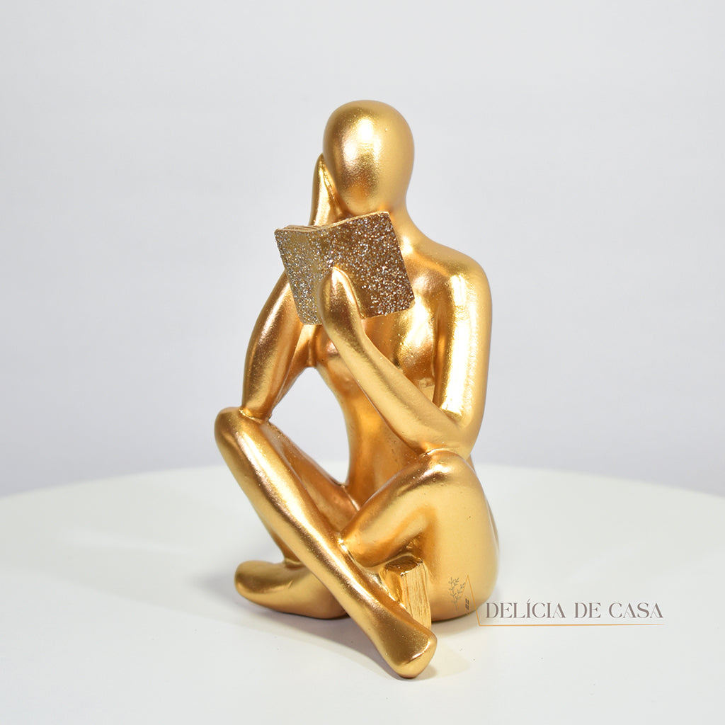 Escultura decorativa dourada de mulher lendo, sentada sobre livros, com detalhes em glitter, ideal para decoração moderna e sofisticada