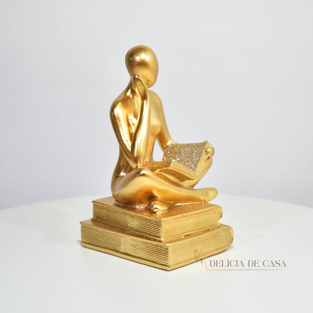 Escultura decorativa dourada de mulher lendo, sentada sobre livros, com detalhes em glitter, ideal para decoração moderna e sofisticada