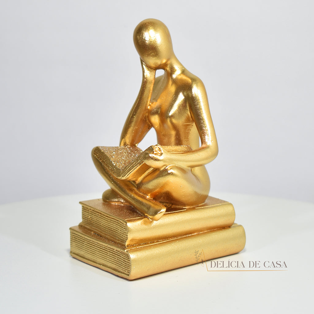 Escultura decorativa dourada de mulher lendo, sentada sobre livros, com detalhes em glitter, ideal para decoração moderna e sofisticada