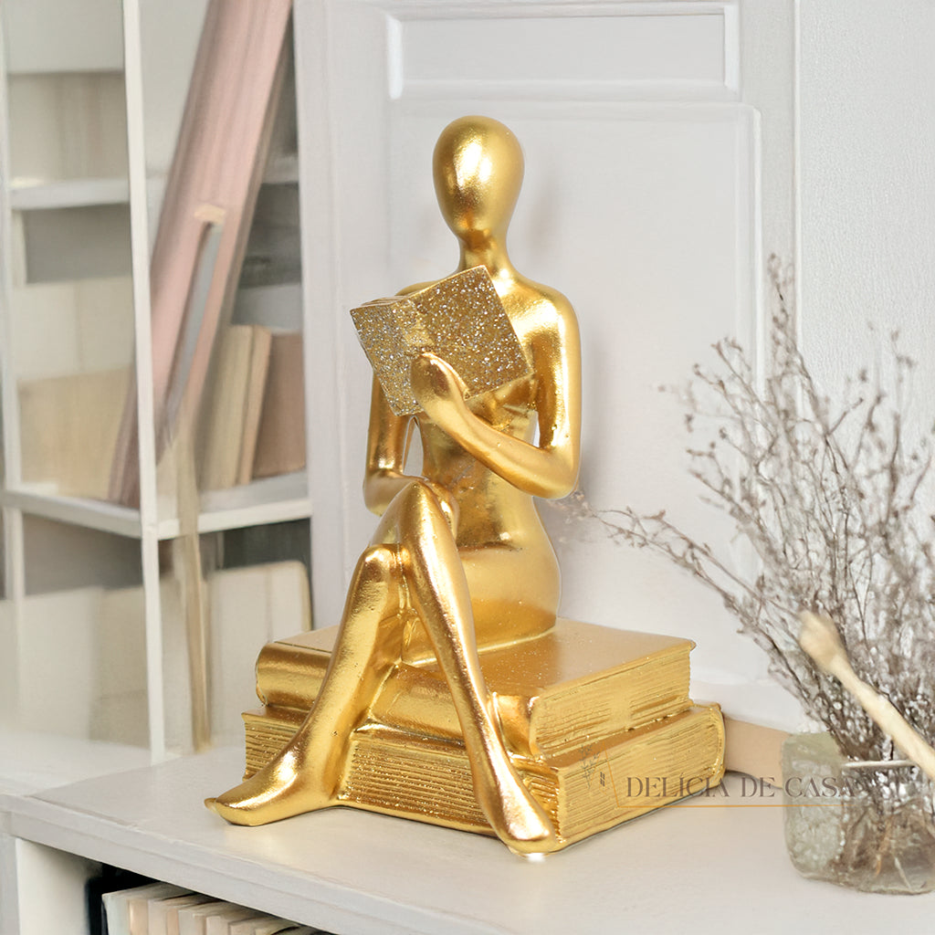 Escultura decorativa dourada de mulher lendo, sentada sobre livros, com detalhes em glitter, ideal para decoração moderna e sofisticada