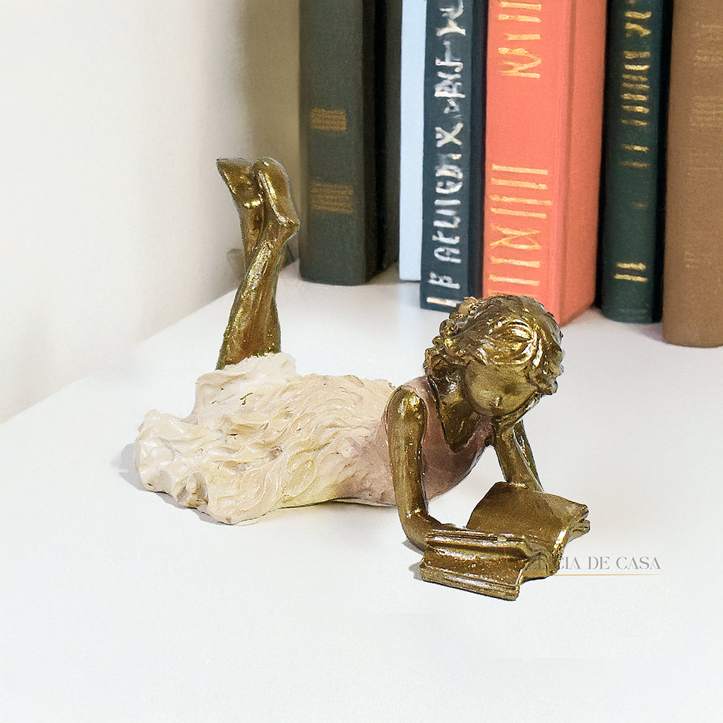 Escultura decorativa de menina lendo, deitada com as pernas cruzadas, feita em resina dourada com vestido em tons claros. Decoração para apaixonados por livros.
