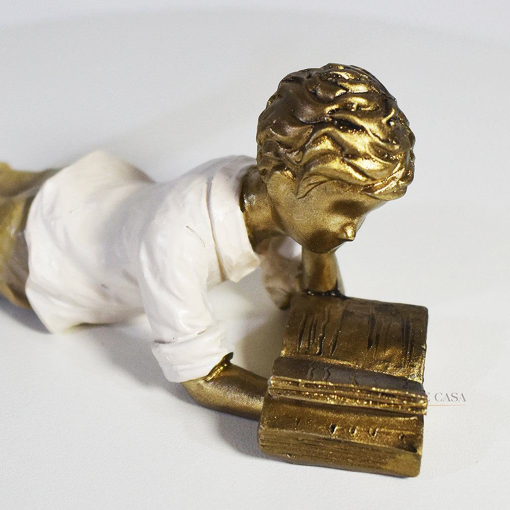 Escultura decorativa de menino lendo, deitada com as pernas cruzadas, feita em resina dourada com vestido em tons claros. Decoração para apaixonados por livros.