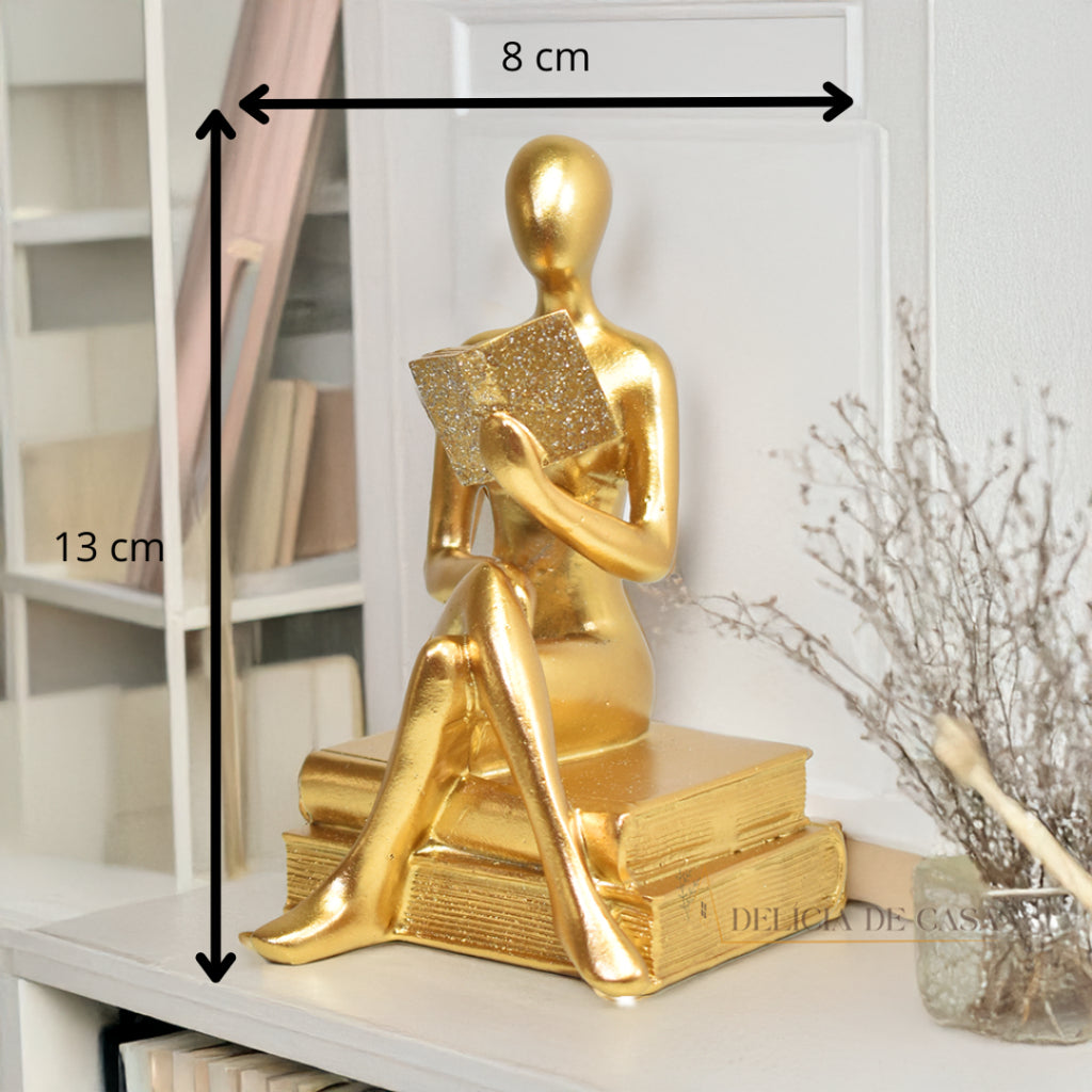 Escultura decorativa dourada de mulher lendo, sentada sobre livros, com detalhes em glitter, ideal para decoração moderna e sofisticada