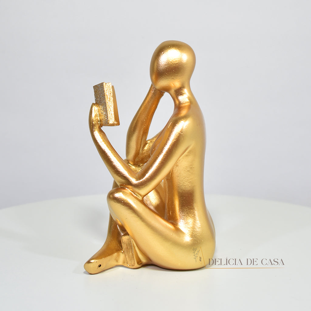 Escultura decorativa dourada de mulher lendo, sentada sobre livros, com detalhes em glitter, ideal para decoração moderna e sofisticada