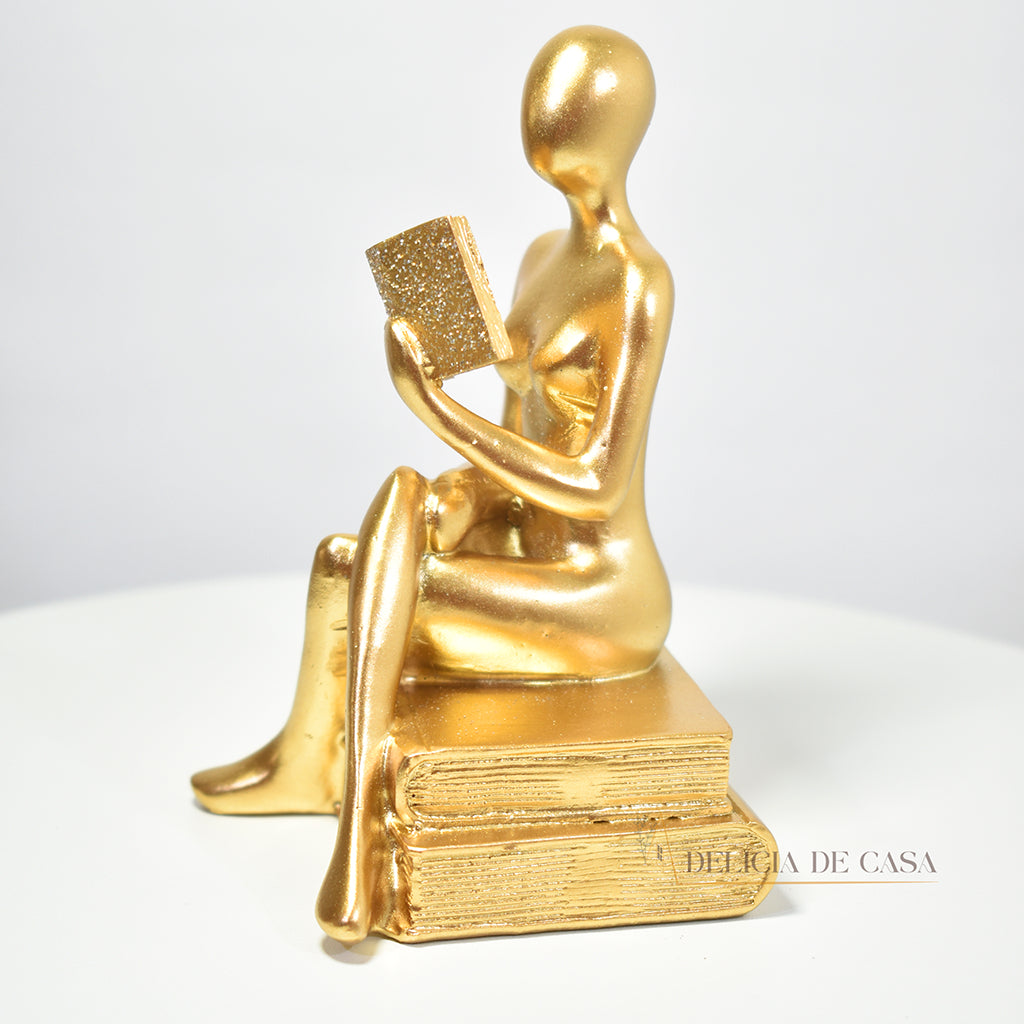 Escultura decorativa dourada de mulher lendo, sentada sobre livros, com detalhes em glitter, ideal para decoração moderna e sofisticada