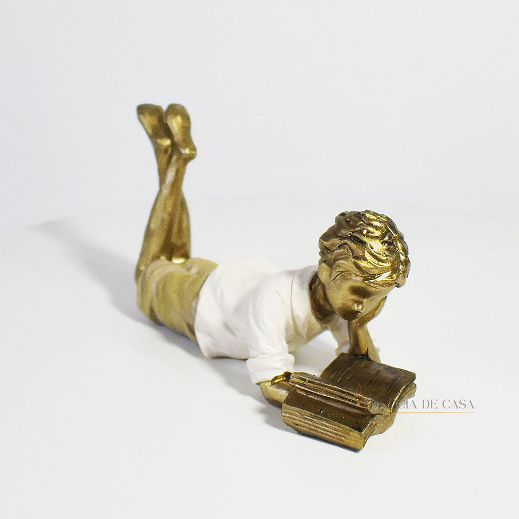 Escultura decorativa de menino lendo, deitada com as pernas cruzadas, feita em resina dourada com vestido em tons claros. Decoração para apaixonados por livros.