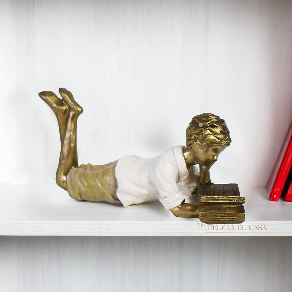 Escultura decorativa de menino lendo, deitada com as pernas cruzadas, feita em resina dourada com vestido em tons claros. Decoração para apaixonados por livros.