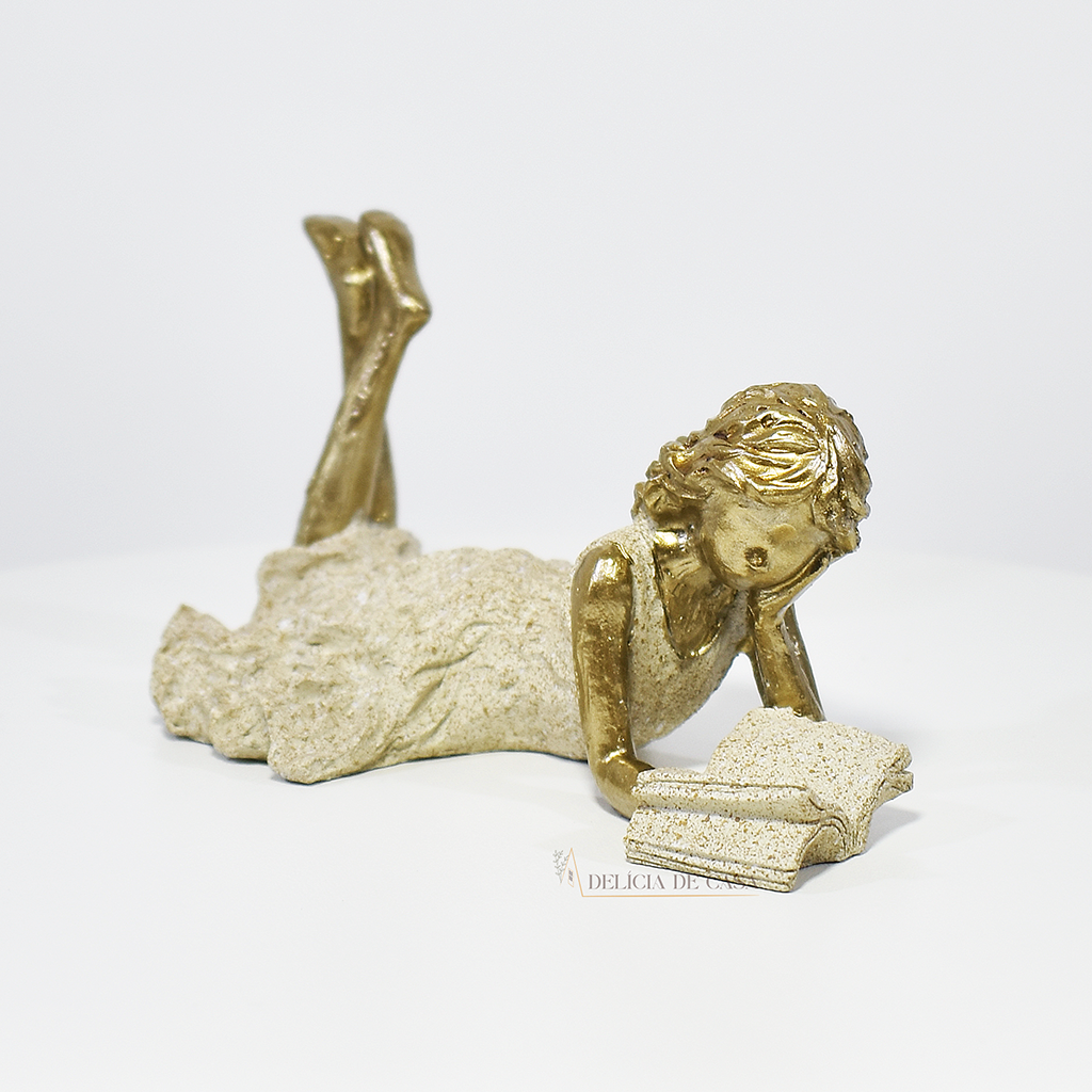Escultura decorativa de menina lendo, deitada com as pernas cruzadas, feita em resina dourada com vestido em tons claros. Decoração para apaixonados por livros.