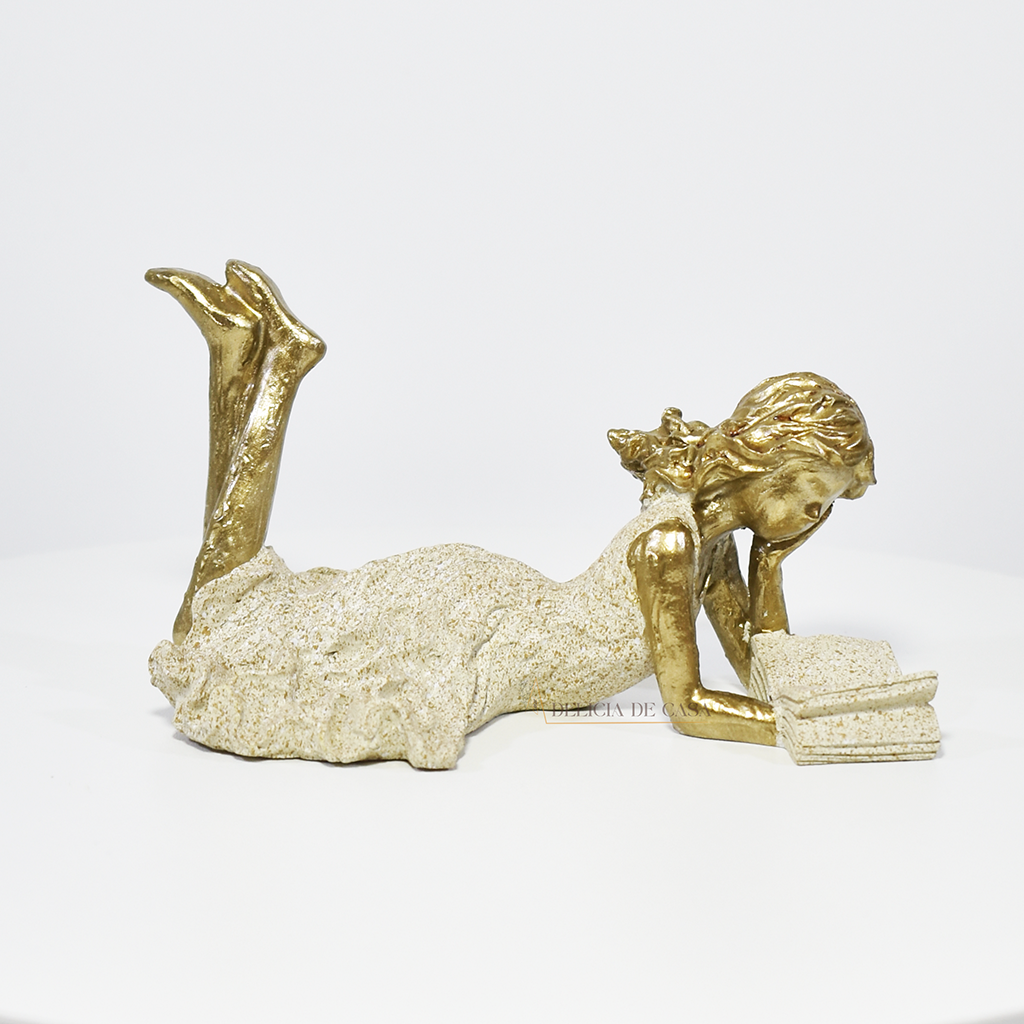 Escultura decorativa de menina lendo, deitada com as pernas cruzadas, feita em resina dourada com vestido em tons claros. Decoração para apaixonados por livros.