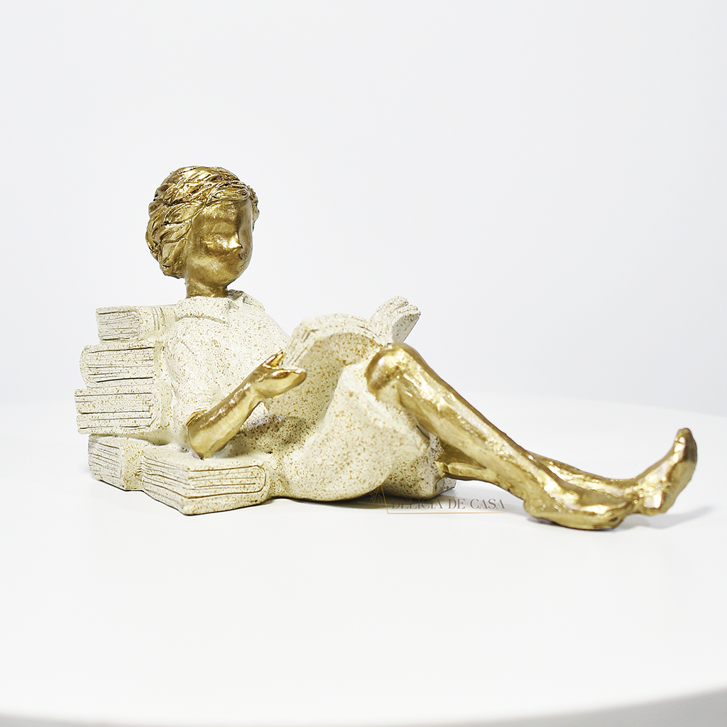 Escultura decorativa de menino lendo, deitada com as pernas cruzadas, feita em resina dourada com vestido em tons claros. Decoração para apaixonados por livros.