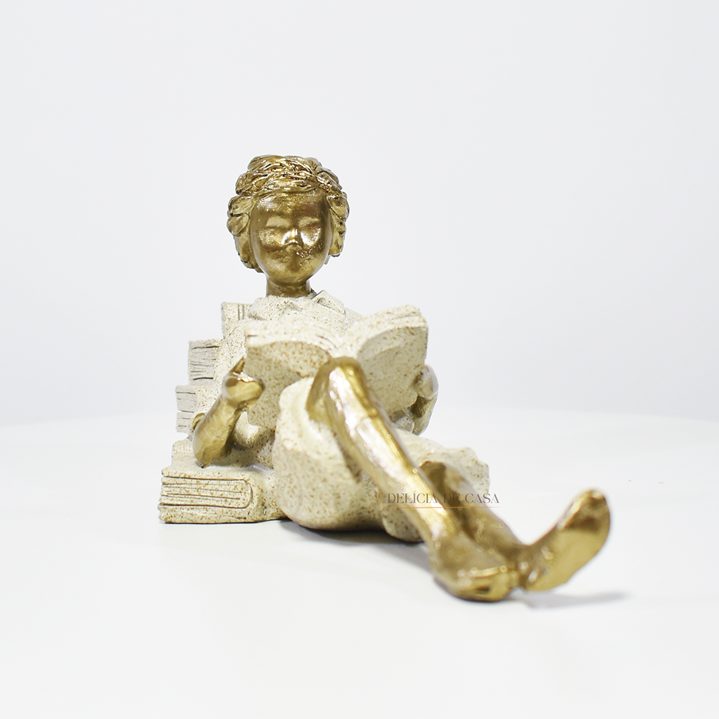 Escultura decorativa de menino lendo, deitada com as pernas cruzadas, feita em resina dourada com vestido em tons claros. Decoração para apaixonados por livros.