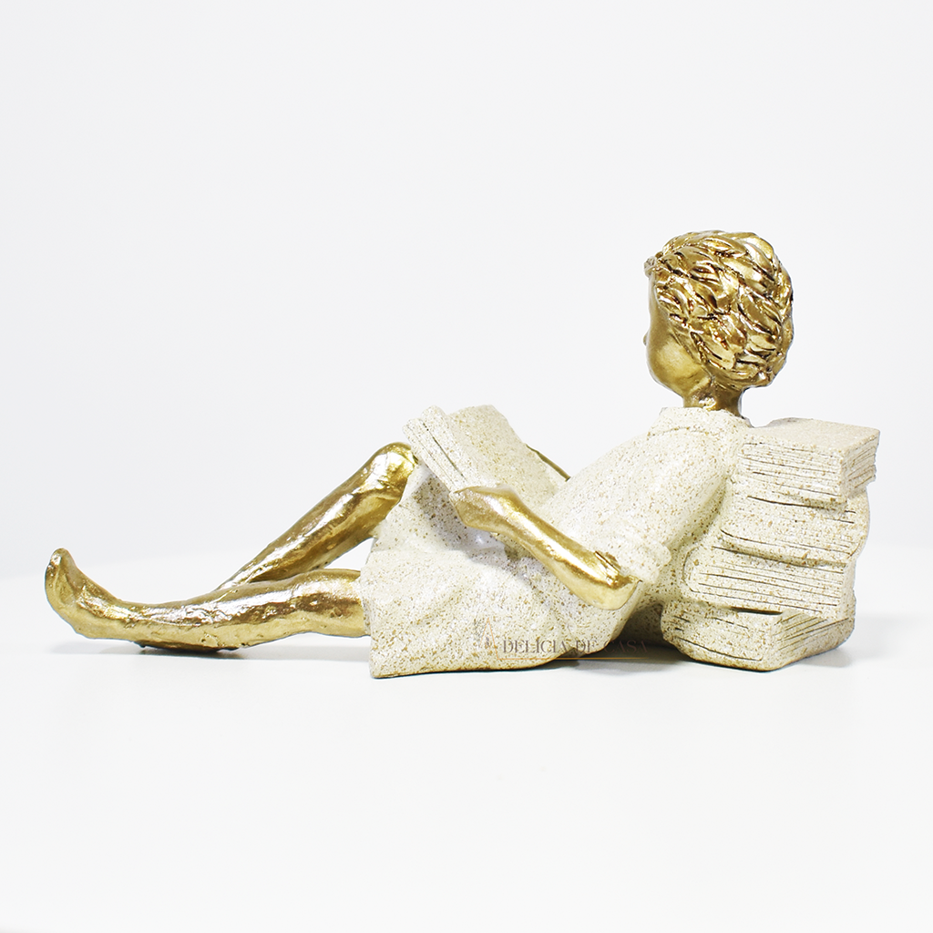 Escultura decorativa de menino lendo, deitada com as pernas cruzadas, feita em resina dourada com vestido em tons claros. Decoração para apaixonados por livros.