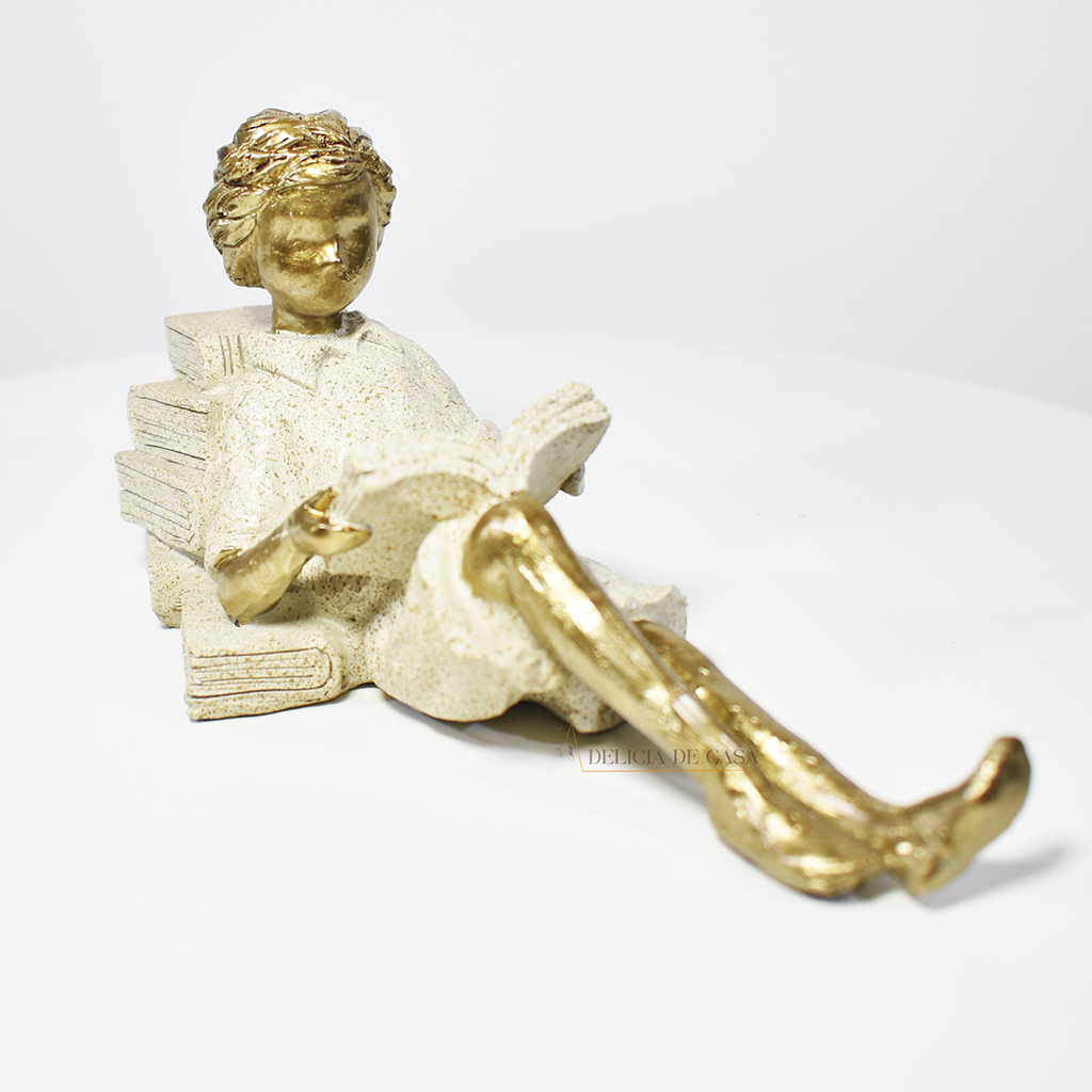 Escultura decorativa de menino lendo, deitada com as pernas cruzadas, feita em resina dourada com vestido em tons claros. Decoração para apaixonados por livros.