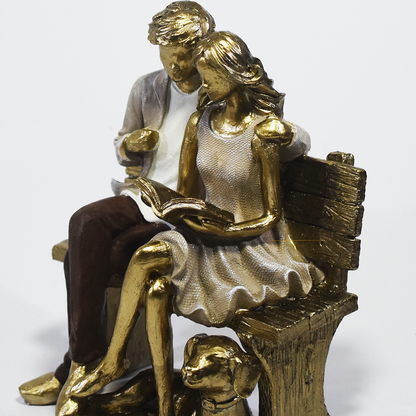 Escultura decorativa em resina de casal sentado em banco lendo um livro, acompanhados por um cachorro aos pés, com acabamento dourado, branco e marrom. Peça elegante para decoração de interiores medindo 15x10x18 cm.