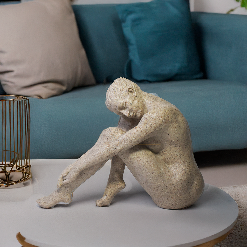 Escultura decorativa feminina em resina com acabamento que simula pedra natural, representando uma figura sentada em posição contemplativa.