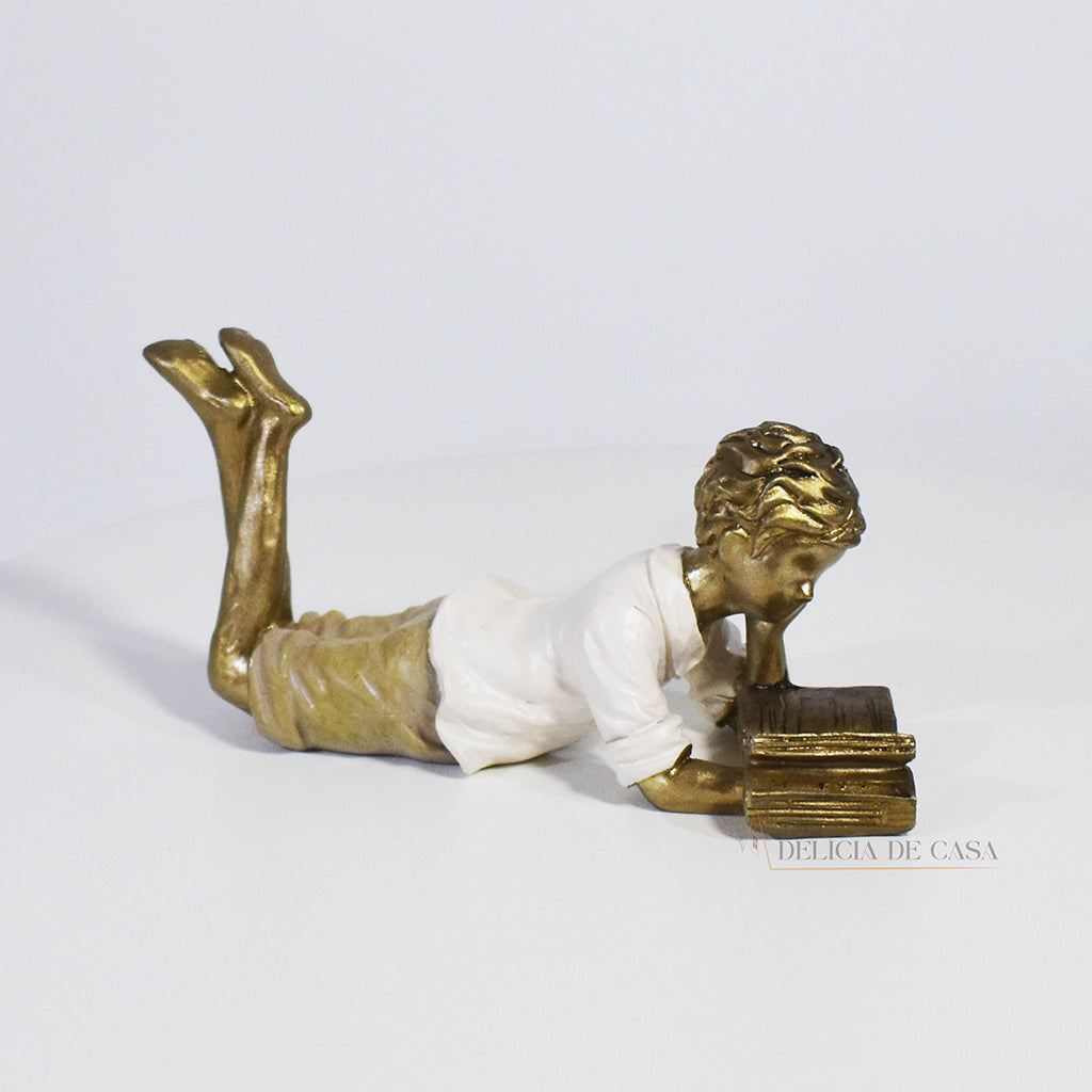 Escultura decorativa de menino lendo, deitada com as pernas cruzadas, feita em resina dourada com vestido em tons claros. Decoração para apaixonados por livros.

