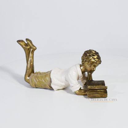 Escultura decorativa de menino lendo, deitada com as pernas cruzadas, feita em resina dourada com vestido em tons claros. Decoração para apaixonados por livros.


