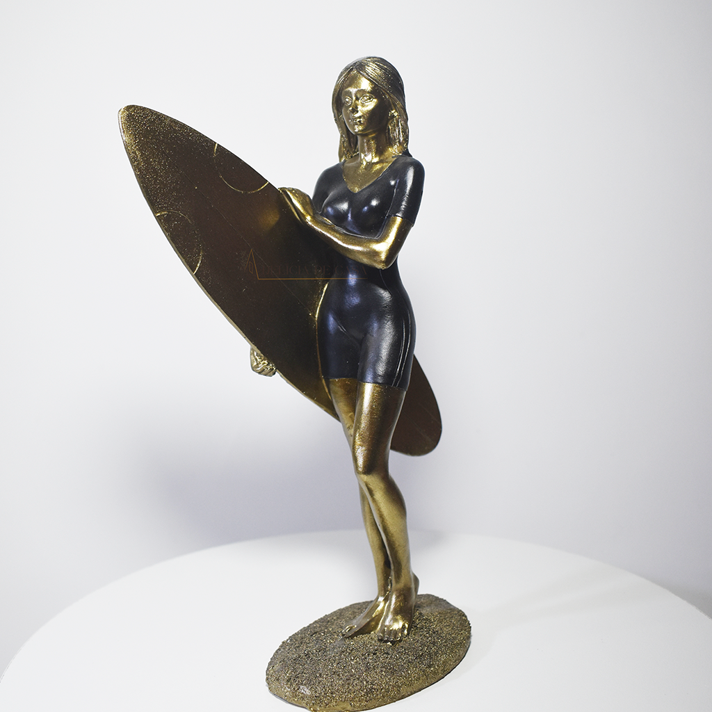 Escultura decorativa em resina de surfista feminina segurando uma prancha de surfe, com acabamento dourado e preto, ideal para decoração moderna e praiana.