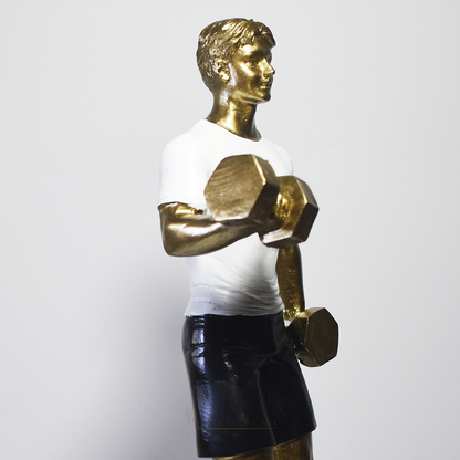 Escultura decorativa em resina representando atleta realizando exercício com halteres, com acabamento dourado, camiseta branca e shorts pretos.