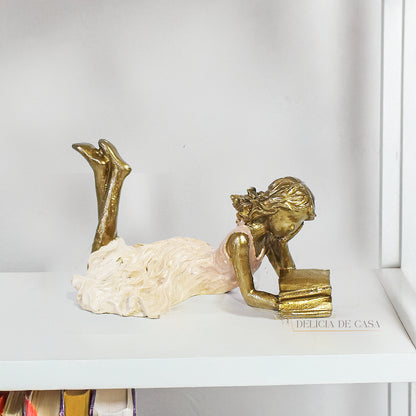 Escultura decorativa de menina lendo, deitada com as pernas cruzadas, feita em resina dourada com vestido em tons claros. Decoração para apaixonados por livros.

