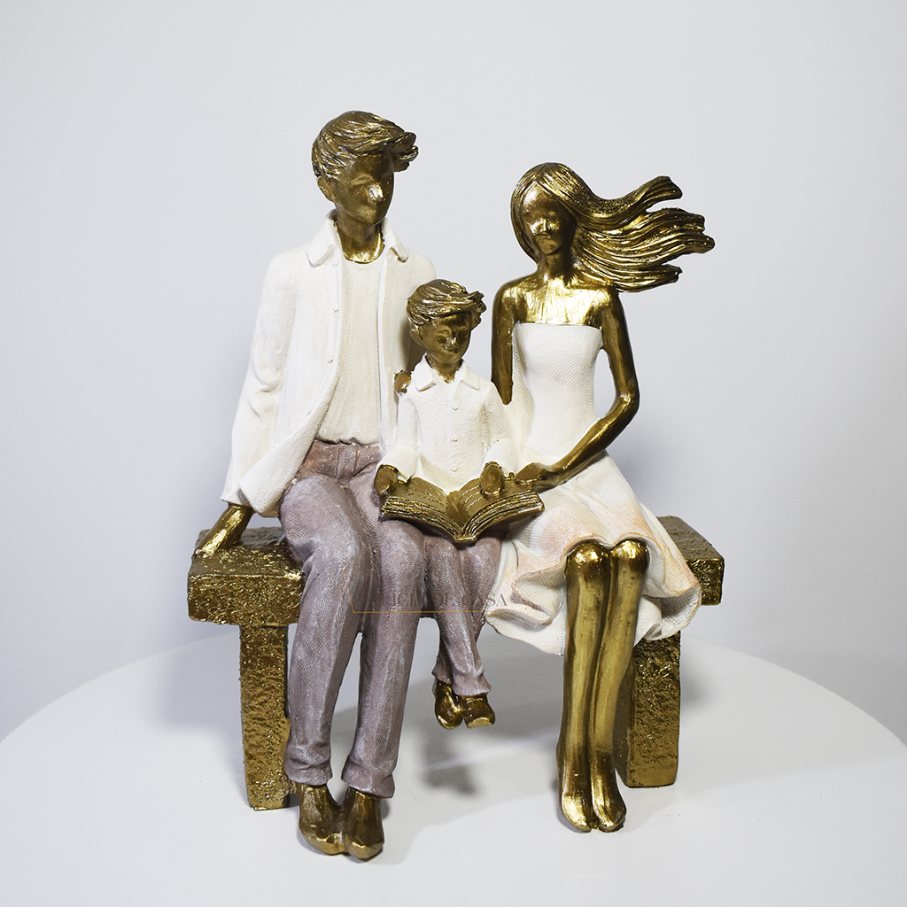Escultura decorativa em resina representando pai, mãe e filho sentados em um banco lendo um livro, simbolizando união familiar e momentos de aprendizado.