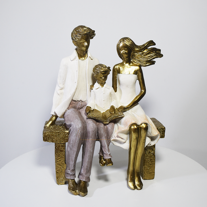 Escultura decorativa em resina representando pai, mãe e filho sentados em um banco lendo um livro, simbolizando união familiar e momentos de aprendizado.