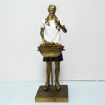 Escultura decorativa em resina de churrasqueiro ao lado de uma churrasqueira, com acabamento dourado, branco e preto.