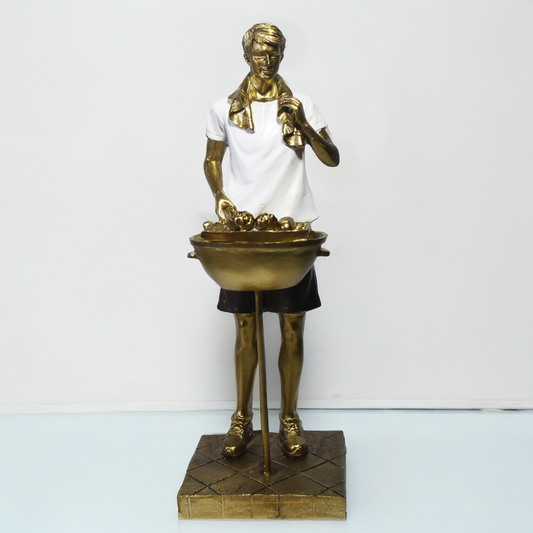 Escultura decorativa em resina de churrasqueiro ao lado de uma churrasqueira, com acabamento dourado, branco e preto.