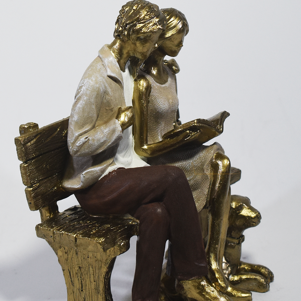 Escultura decorativa em resina de casal sentado em banco lendo um livro, acompanhados por um cachorro aos pés, com acabamento dourado, branco e marrom. Peça elegante para decoração de interiores medindo 15x10x18 cm.