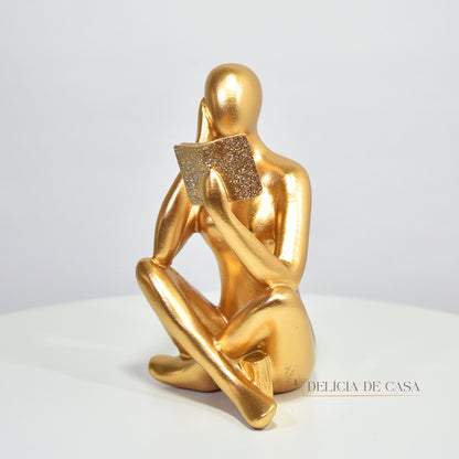 Escultura decorativa dourada de mulher lendo, sentada sobre livros, com detalhes em glitter, ideal para decoração moderna e sofisticada