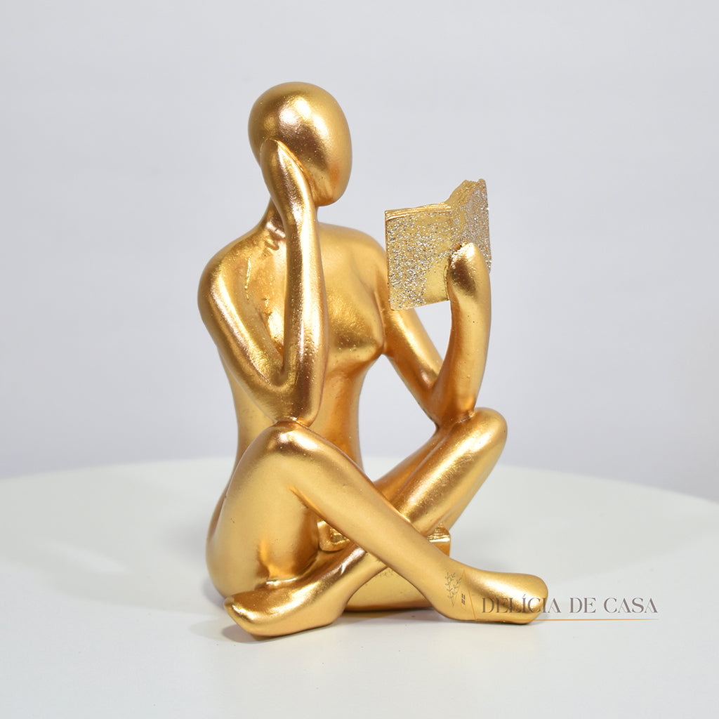 Escultura decorativa dourada de mulher lendo, sentada sobre livros, com detalhes em glitter, ideal para decoração moderna e sofisticada