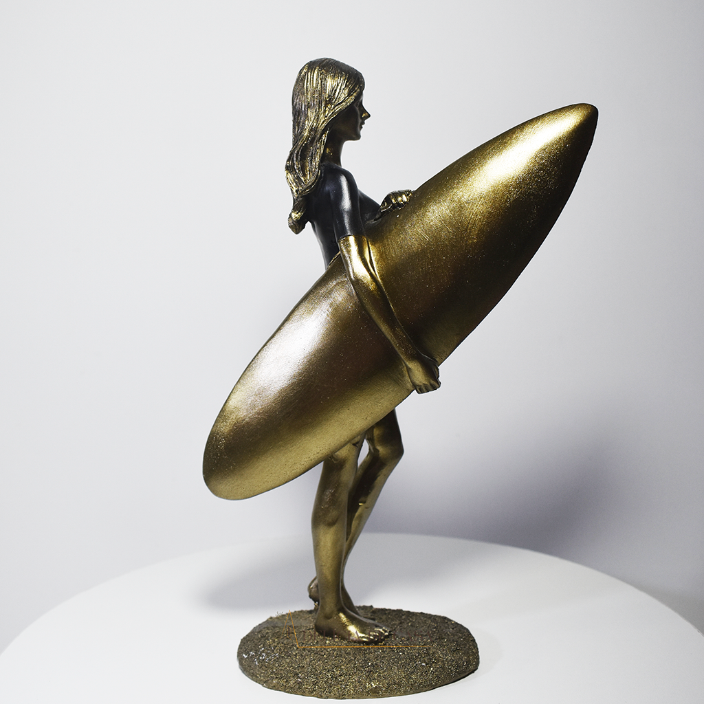 Escultura decorativa em resina de surfista feminina segurando uma prancha de surfe, com acabamento dourado e preto, ideal para decoração moderna e praiana.