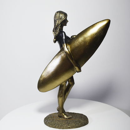 Escultura decorativa em resina de surfista feminina segurando uma prancha de surfe, com acabamento dourado e preto, ideal para decoração moderna e praiana.