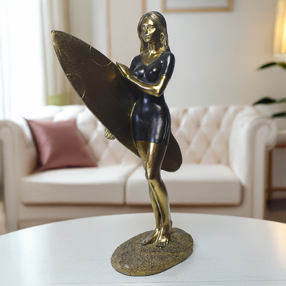 Escultura decorativa em resina de surfista feminina segurando uma prancha de surfe, com acabamento dourado e preto, ideal para decoração moderna e praiana.