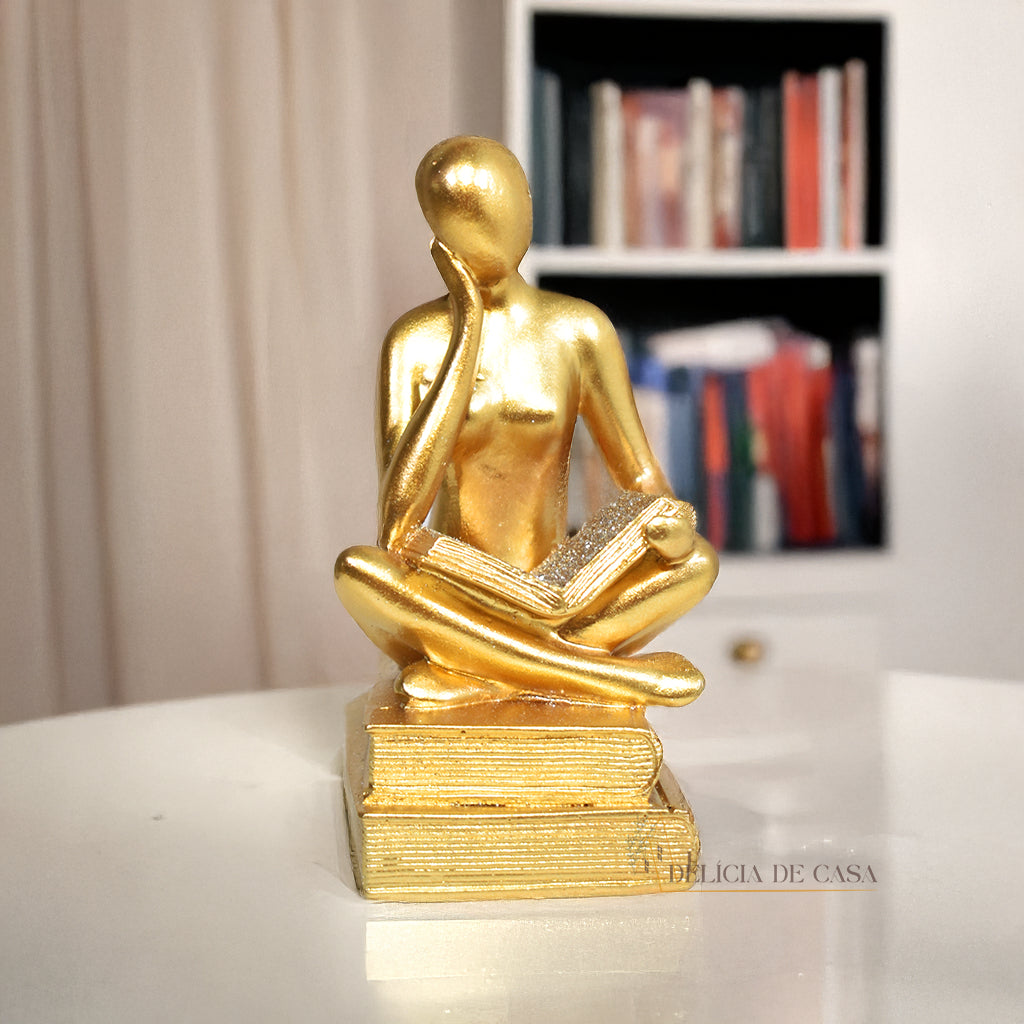 Escultura decorativa dourada de mulher lendo, sentada sobre livros, com detalhes em glitter, ideal para decoração moderna e sofisticada