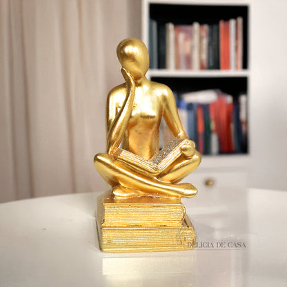 Escultura decorativa dourada de mulher lendo, sentada sobre livros, com detalhes em glitter, ideal para decoração moderna e sofisticada