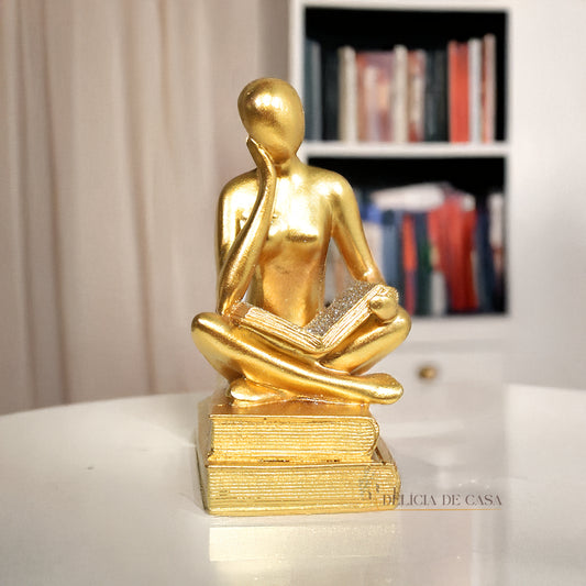 Escultura decorativa dourada de mulher lendo, sentada sobre livros, com detalhes em glitter, ideal para decoração moderna e sofisticada