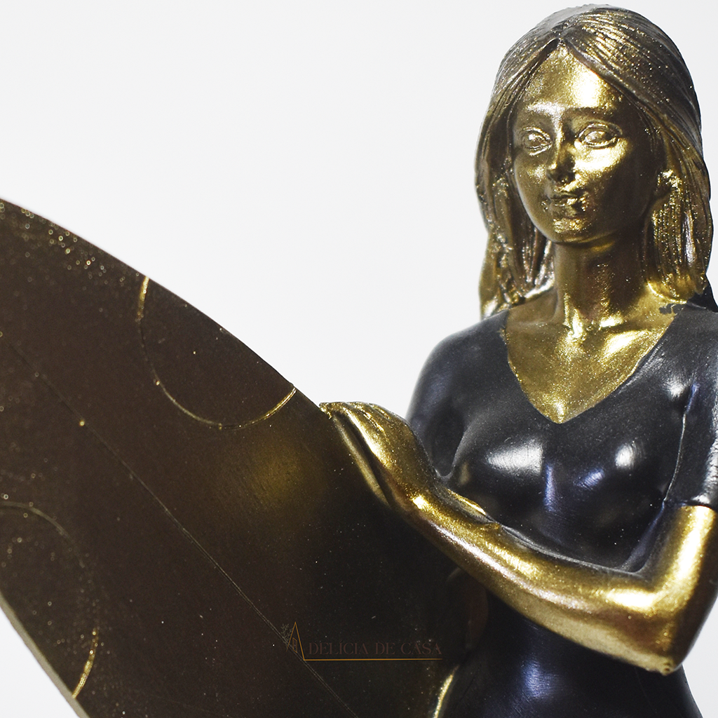 Escultura decorativa em resina de surfista feminina segurando uma prancha de surfe, com acabamento dourado e preto, ideal para decoração moderna e praiana.