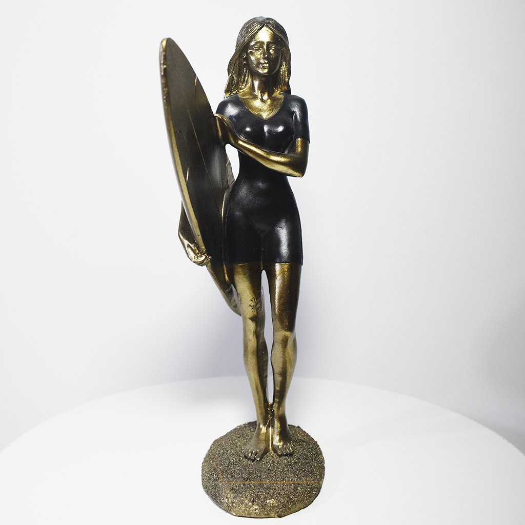 Escultura decorativa em resina de surfista feminina segurando uma prancha de surfe, com acabamento dourado e preto, ideal para decoração moderna e praiana.