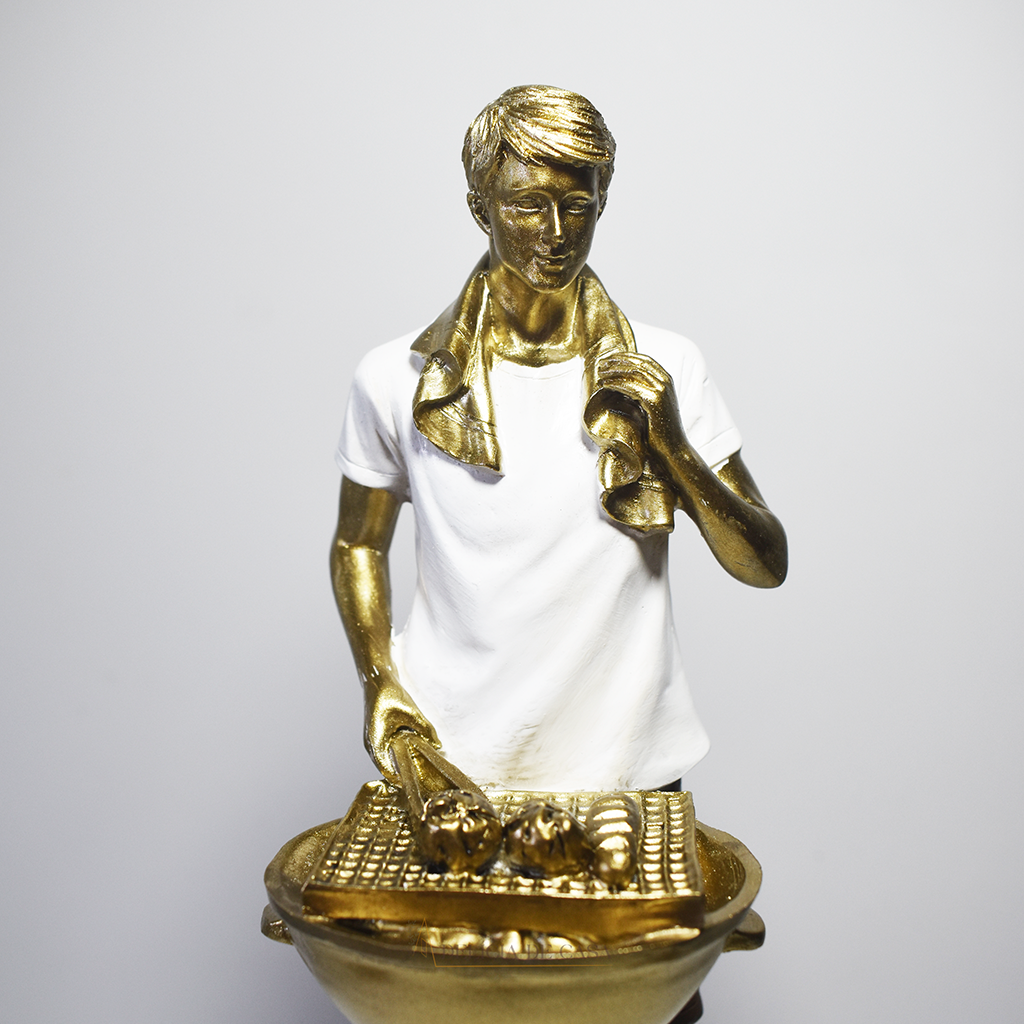 Escultura decorativa em resina de churrasqueiro ao lado de uma churrasqueira, com acabamento dourado, branco e preto.