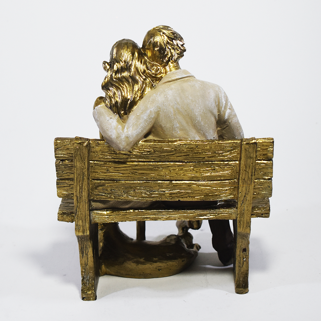 Escultura decorativa em resina de casal sentado em banco lendo um livro, acompanhados por um cachorro aos pés, com acabamento dourado, branco e marrom. Peça elegante para decoração de interiores medindo 15x10x18 cm.