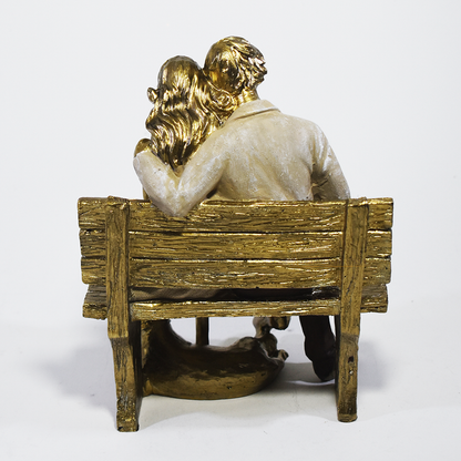 Escultura decorativa em resina de casal sentado em banco lendo um livro, acompanhados por um cachorro aos pés, com acabamento dourado, branco e marrom. Peça elegante para decoração de interiores medindo 15x10x18 cm.