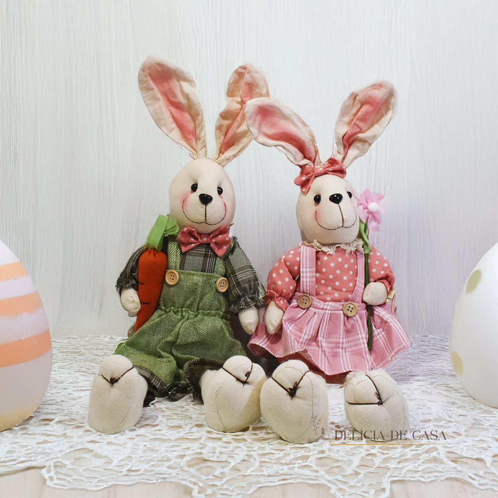 Dois coelhos de pelúcia sentados sobre um móvel, um vestindo macacão verde e segurando uma cenoura, e o outro com um vestido floral, ideal para decoração de Páscoa.