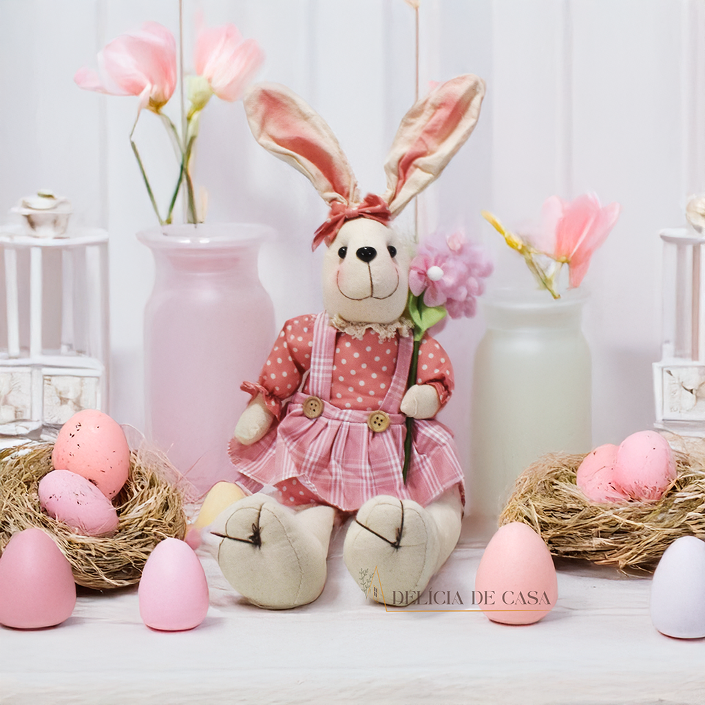 Dois coelhos de pelúcia sentados sobre um móvel, um vestindo macacão verde e segurando uma cenoura, e o outro com um vestido floral, ideal para decoração de Páscoa.